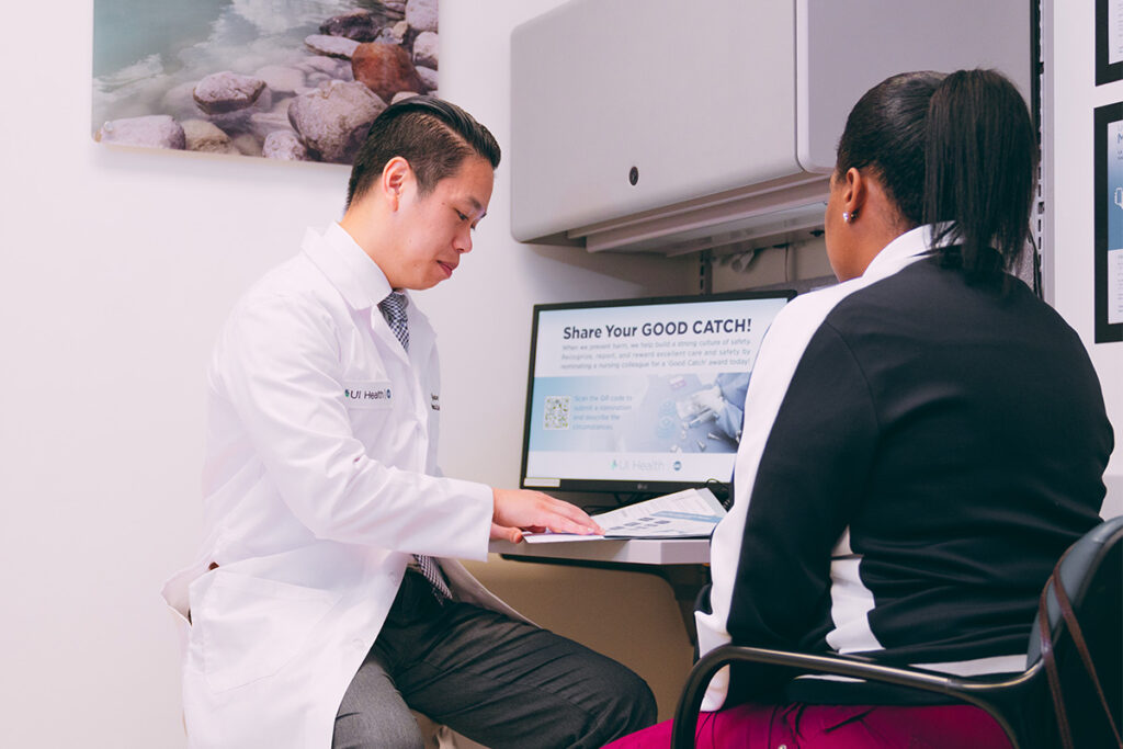 A male doctor in a white coat seated next to a female patient, reviewing papers on a desk.