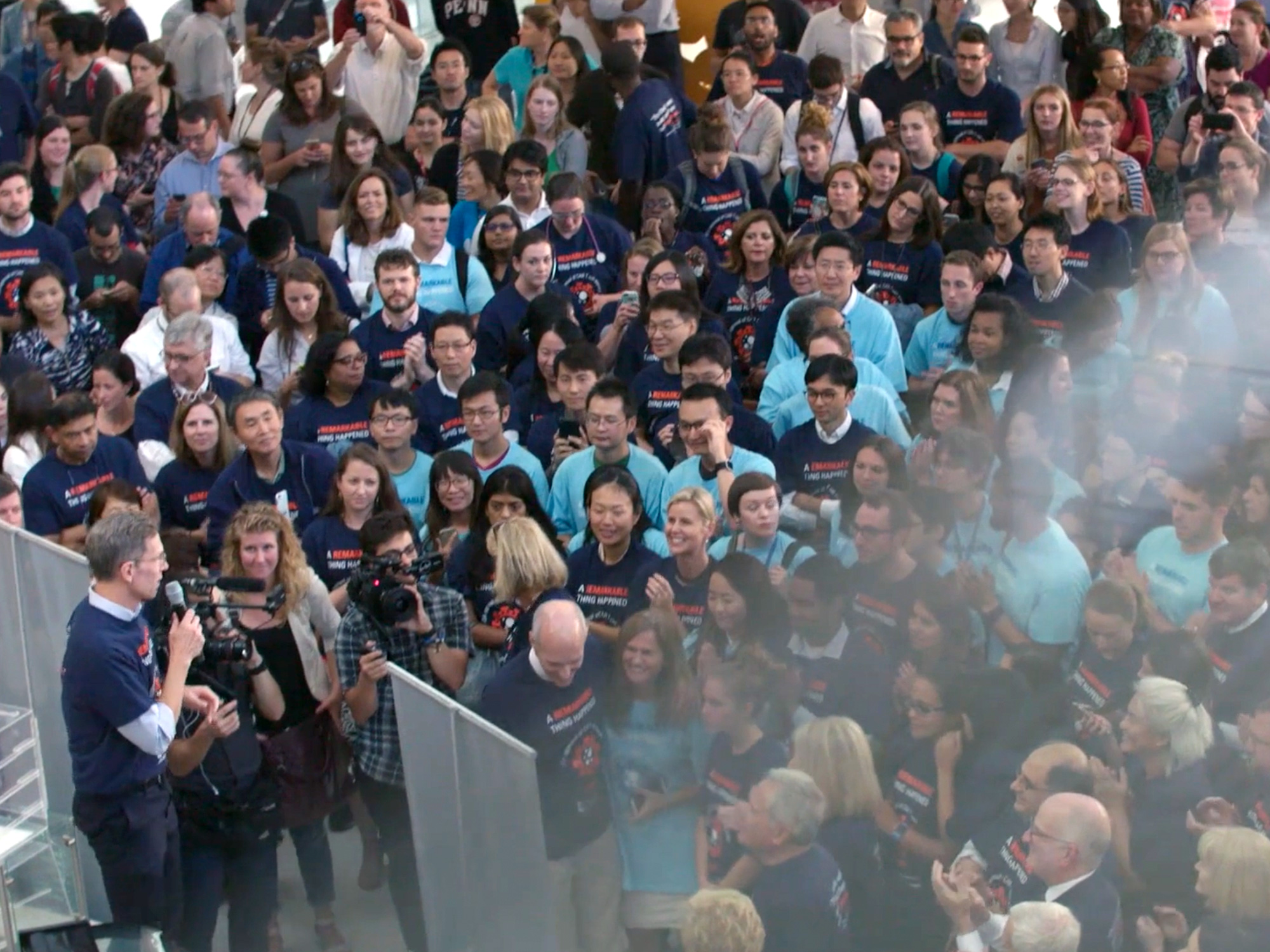 A man with a microphone, at left, addresses a crowd of people wearing Penn Immunotherapy T-shirts.